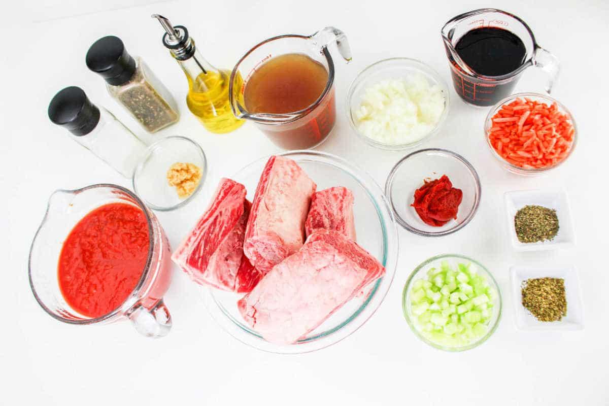 A collection of ingredients for a recipe arranged on a white surface, including raw beef short ribs perfect for Short Rib Tacos, tomato sauce, beef broth, diced onions, shredded carrots, chopped celery, red wine, tomato paste, minced garlic, cooking oil, salt, pepper and dried herbs.