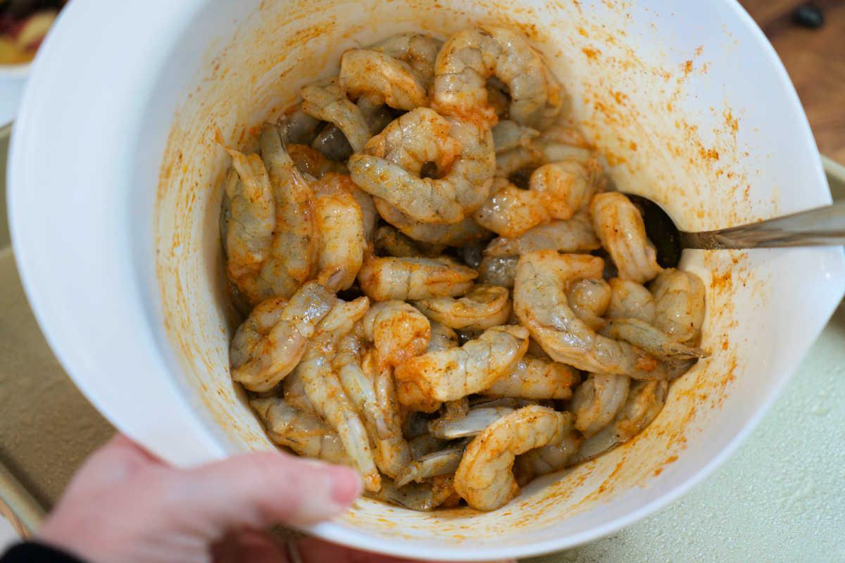 A bowl of raw shrimp and potatoes marinated with spices, being mixed with a spoon.