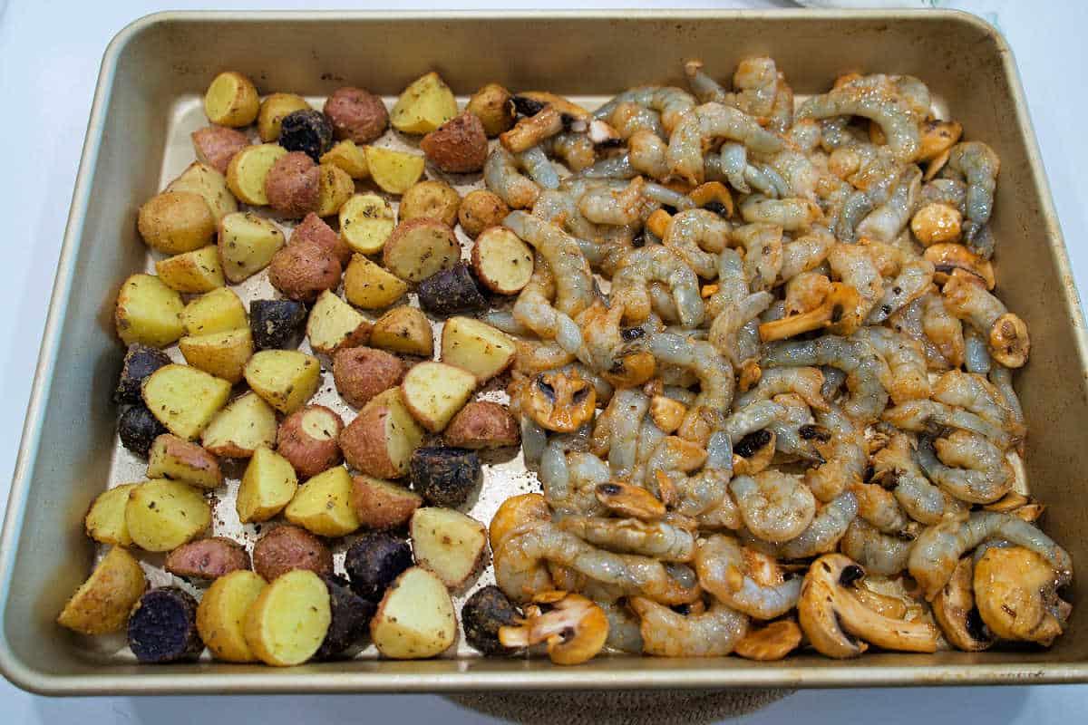 Sheet pan with seasoned baby potatoes on the left and raw shrimp and mushrooms on the right, this shrimp and potatoes combo is ready to bake.