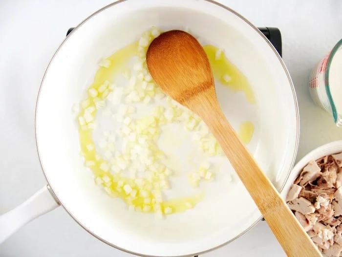 Diced onions and oil sizzle in a white pan with a wooden spoon, next to a bowl of shredded meat—perfect for starting your simple chicken pot pie recipe.