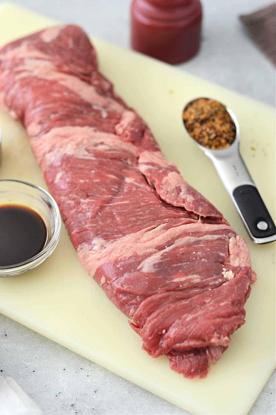 A raw skirt steak rests on the cutting board, ready to become a masterpiece. Alongside it, a bowl of rich soy sauce and a spoonful of spices await their turn to enhance flavors. Imagine preparing skirt steak in the oven with this selection for an unforgettable culinary experience.