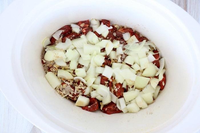 Chopped onions and spices mingle with slow cooker beef tips in a white slow cooker.