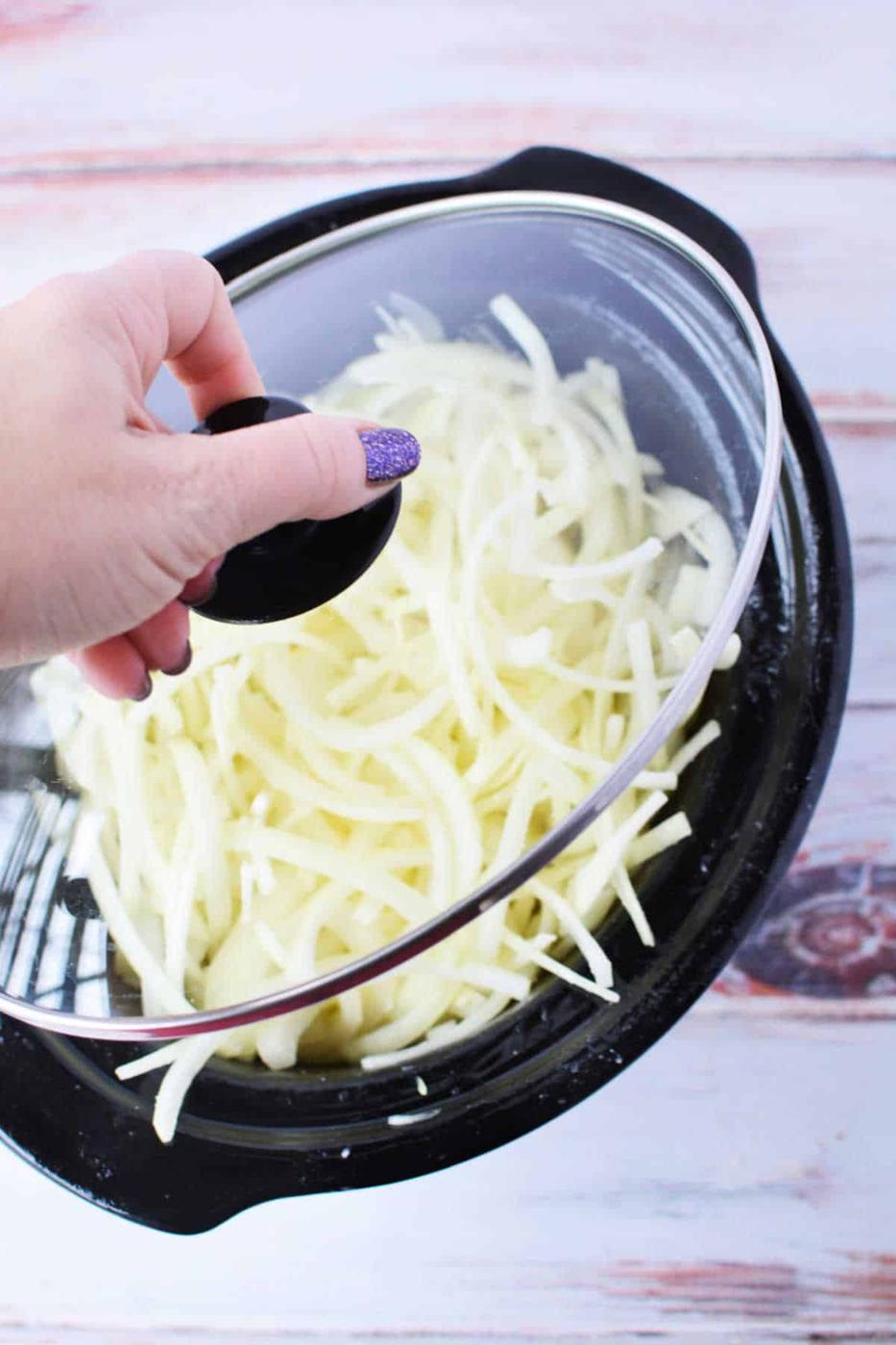 Hand lifting lid from slow cooker filled with shredded potatoes and caramelized onions.