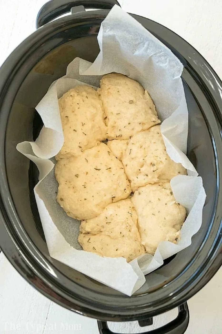 Unbaked herb biscuits, resembling Slow Cooker Rolls, are placed on parchment paper inside a black slow cooker, ready to be cooked.