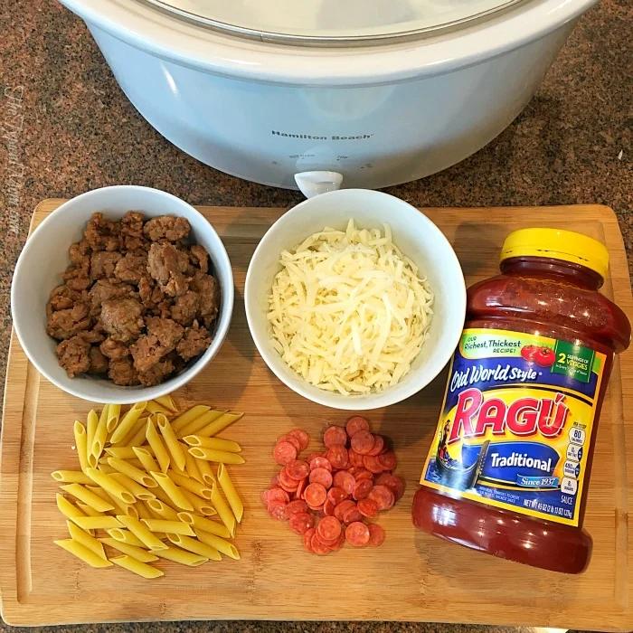Ingredients for a hearty slow cooker pizza casserole are spread across the cutting board: uncooked penne, cooked sausage, shredded cheese, mini pepperoni, and a jar of Ragú sauce, all ready to be combined in the Hamilton Beach slow cooker.