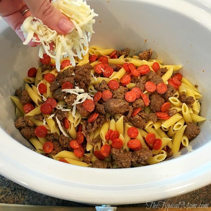 A hand sprinkles cheese over a slow cooker pizza casserole filled with penne pasta, sausage, and pepperoni.
