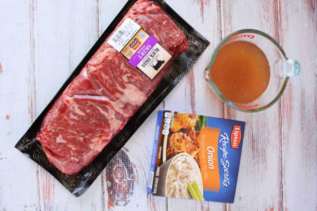 Raw beef steak in packaging, a measuring cup of broth, and a box of onion soup mix on a white wooden surface—perfect ingredients for an easy Slow Cooker Skirt Steak recipe.