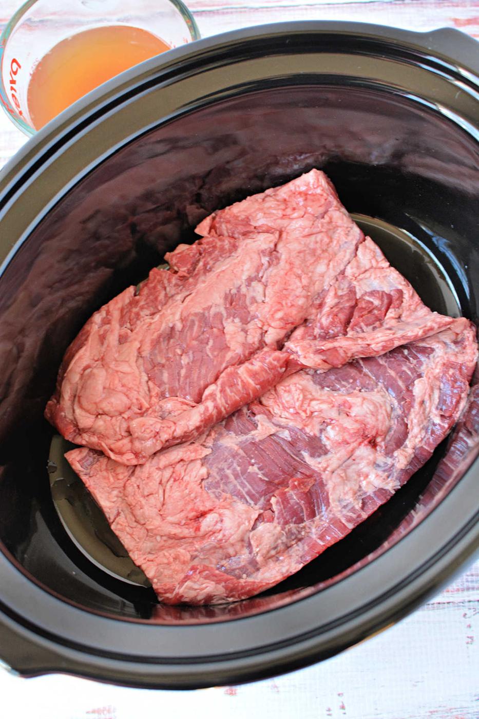 Two raw pieces of beef, ideal for slow cooker skirt steak, are placed inside a black slow cooker, with broth in a measuring cup nearby—perfect for those seeking delicious skirt steak recipes.