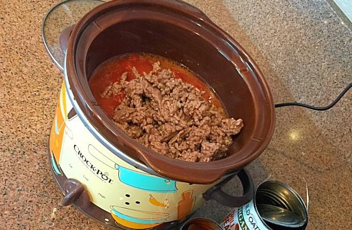 Ground beef and tomato sauce simmering in a Crock-Pot, reminiscent of slow cooker soup recipes, with empty cans scattered on the kitchen countertop.