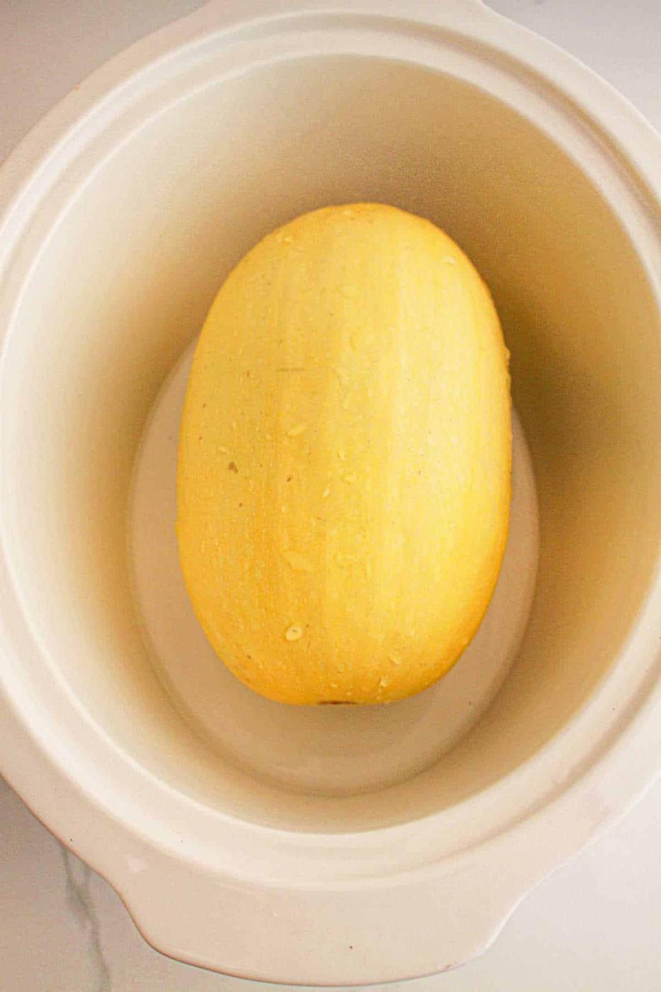 A whole slow cooker spaghetti squash nestles perfectly inside a white slow cooker, ready to transform into a delectable dish.