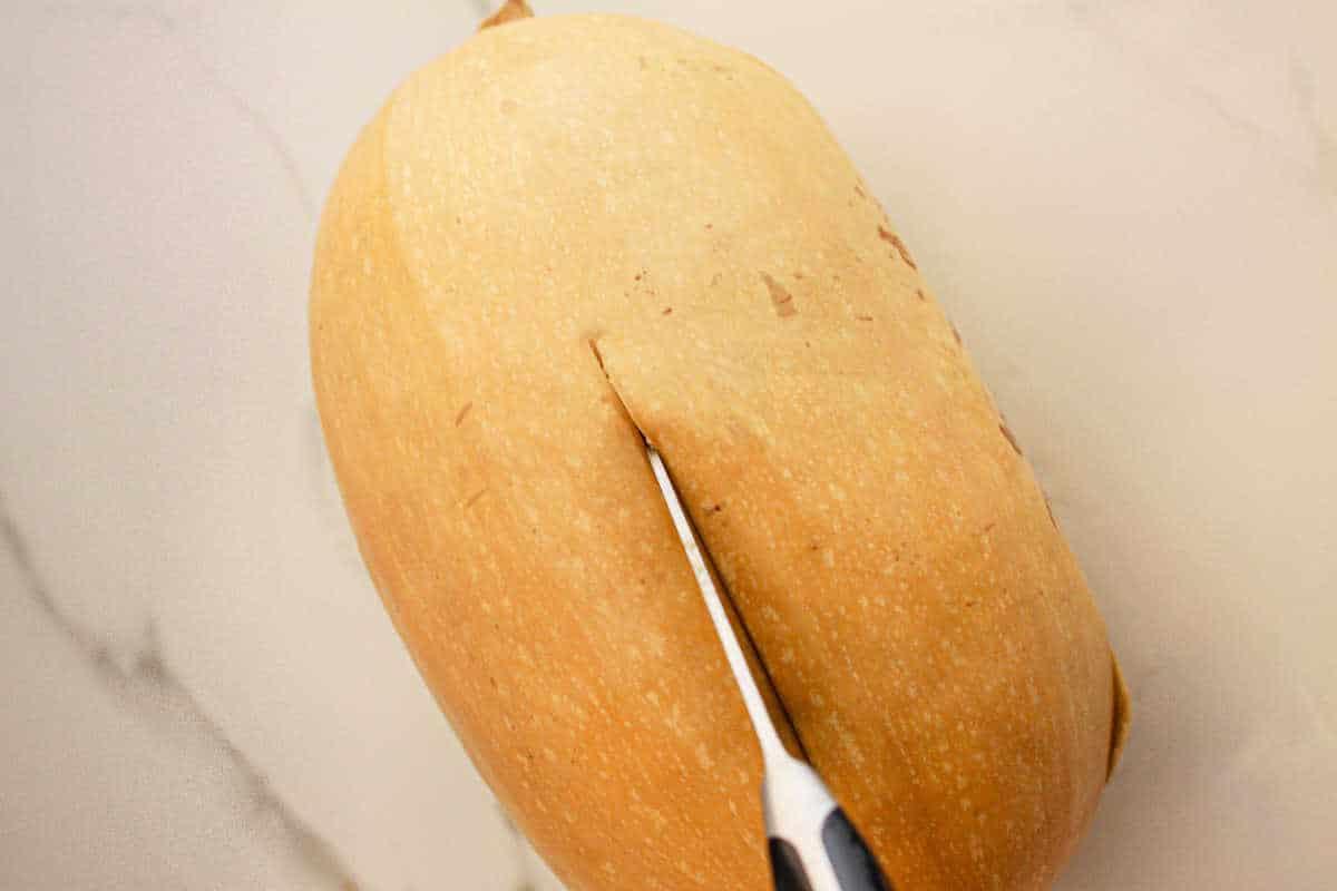 A butternut squash being sliced with a knife on a light-colored countertop, ready to transform into a delicious slow cooker spaghetti squash dish.