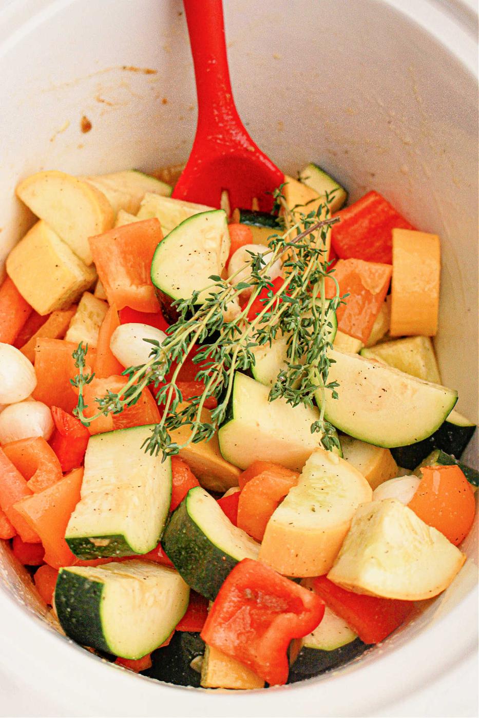 A red spoon rests in a pot filled with chopped fresh vegetables, including zucchini, bell peppers, and onions. A sprig of thyme is placed on top, adding a touch of green to the mix. The seasoned vegetables appear ready for cooking in a slow cooker.