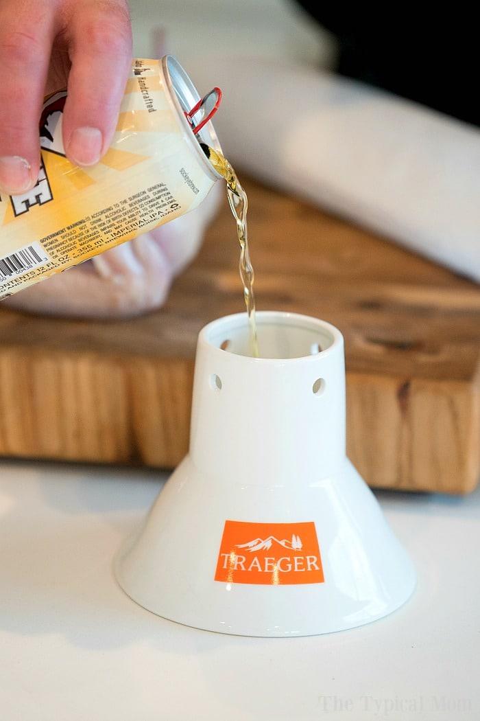 A person skillfully pours beer into a white Traeger chicken throne on a wooden countertop, preparing the perfect base for a delicious smoked beer can chicken.