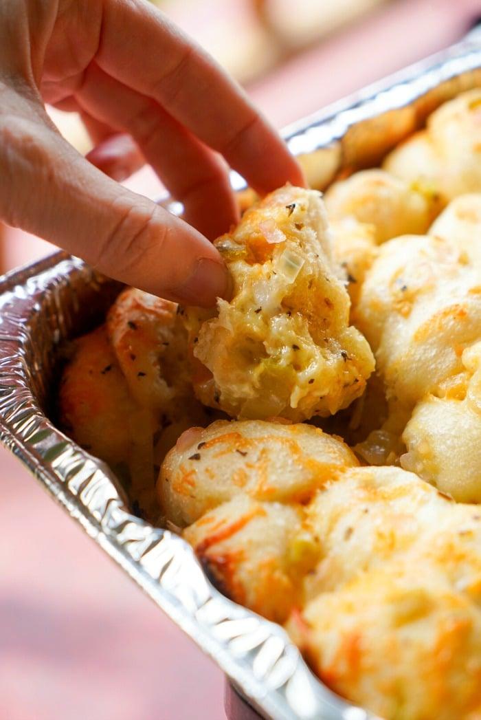 A hand picks up a piece of smoked, cheesy baked bread from a foil tray filled with golden, fluffy rolls. The bread appears slightly crispy with a rich, melted cheese topping.