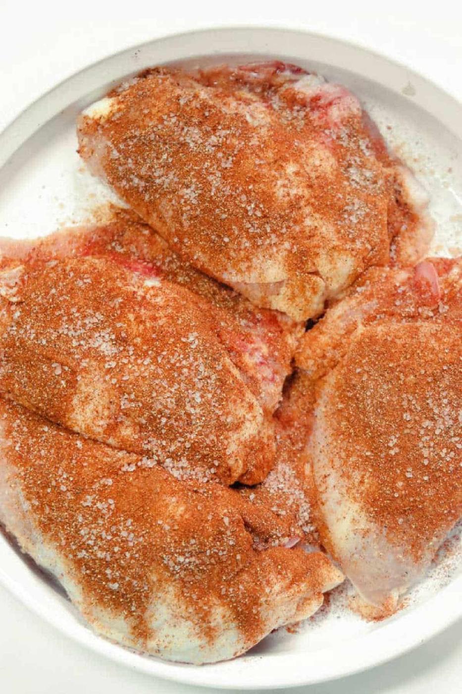A plate with raw chicken pieces seasoned with a generous amount of dry rub spices, including salt and a reddish spice mix, ready for cooking as smoked chicken breast. The seasoning is evenly distributed over the chicken pieces, which are placed closely together.