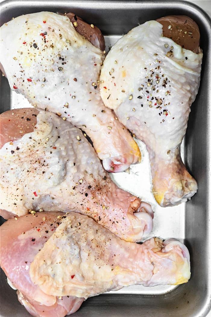 Four raw chicken legs in a metal baking dish, seasoned with salt, pepper, and spices, ready to transform into smoky chicken drumsticks.
