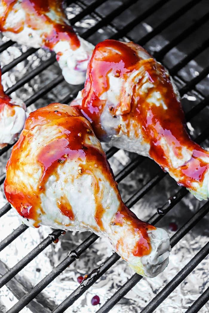 Smoked chicken drumsticks brushed with red sauce sizzle on the grill. Foil beneath catches drips as the glossy glaze coats each piece, enhancing their mouthwatering appeal.