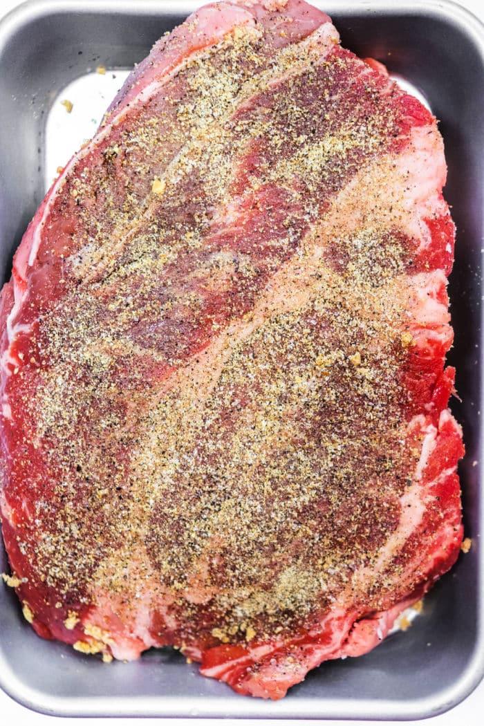 Raw seasoned beef brisket in a metal baking pan, coated with pepper and spices, ready for cooking—perfect for those who love the flavors of a classic smoked chuck roast.