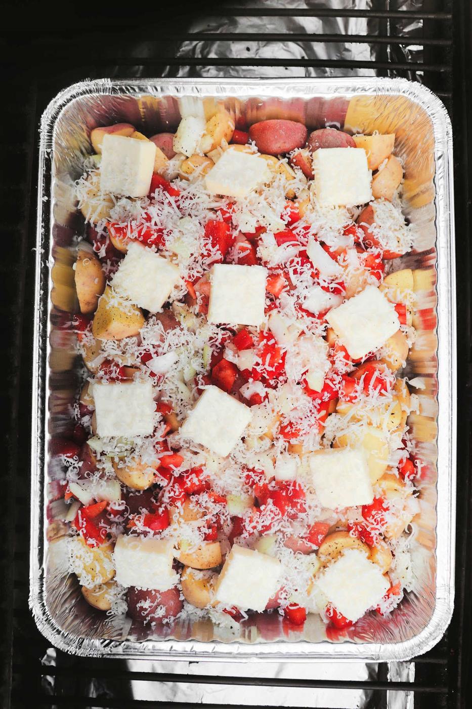 Foil pan filled with chopped vegetables, grated cheese, and cubed butter, perfect for making smoked cheesy potatoes and ready to be cooked.