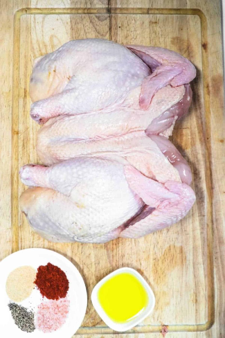 Raw smoked Spatchcock Chicken on a cutting board, with spices and olive oil in small dishes beside it.