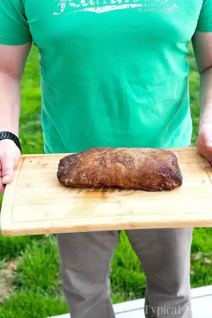 A person stands on the lush grass, holding a wooden board showcasing a perfectly smoked boneless pork loin.