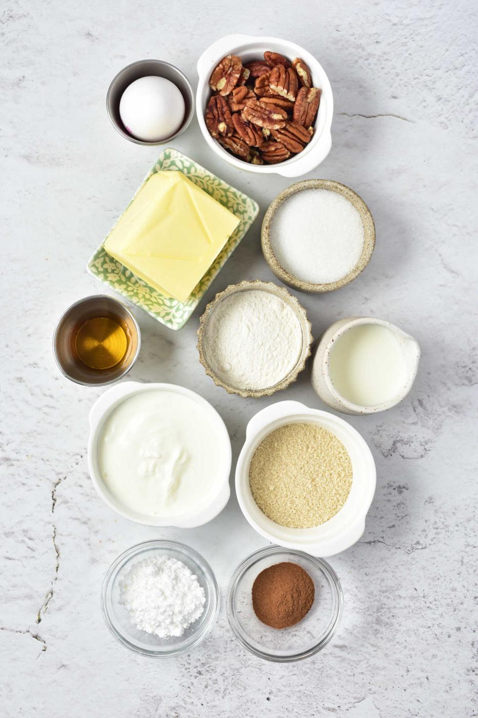 Ingredients laid out on a surface for a Sock it to me cake recipe with cake mix: egg, pecans, butter, sugar, flour, milk, vanilla, sour cream, powdered sugar, cinnamon.