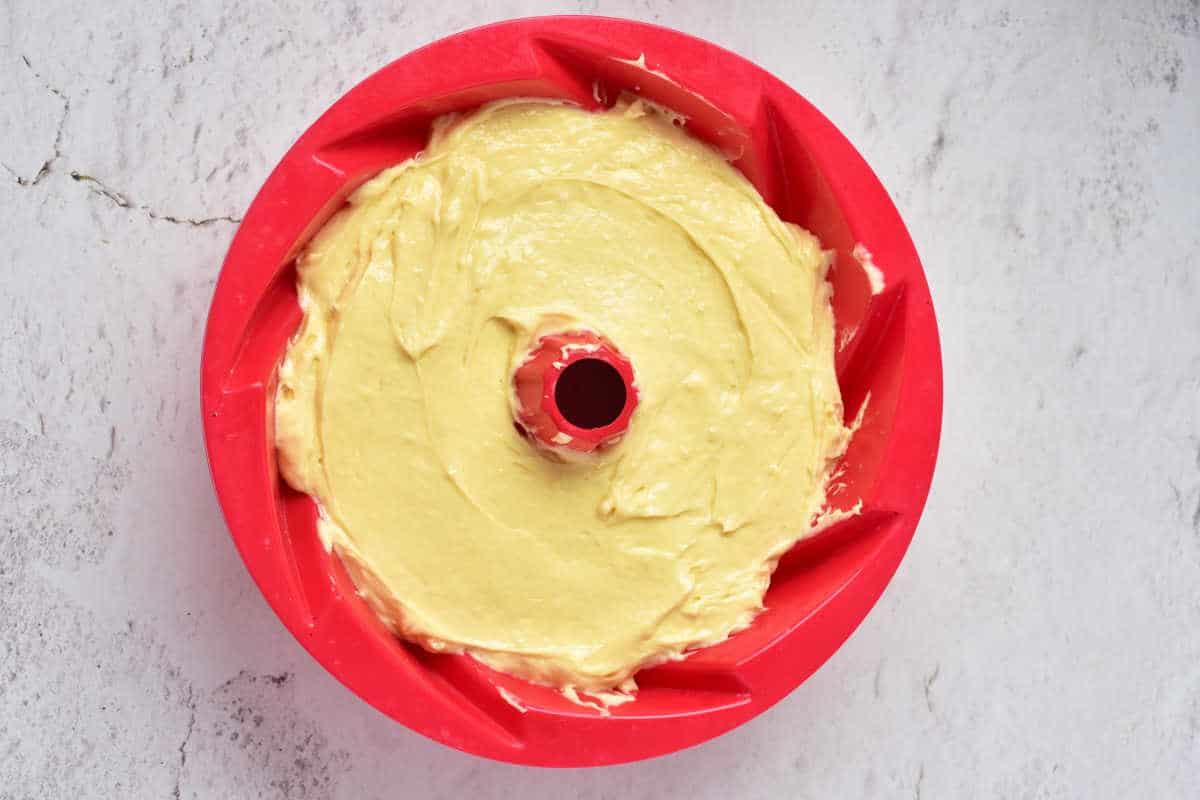 A red bundt pan filled with yellow cake batter sits invitingly on a light gray surface, ready to transform into a delightful Sock it to me cake using a convenient cake mix.