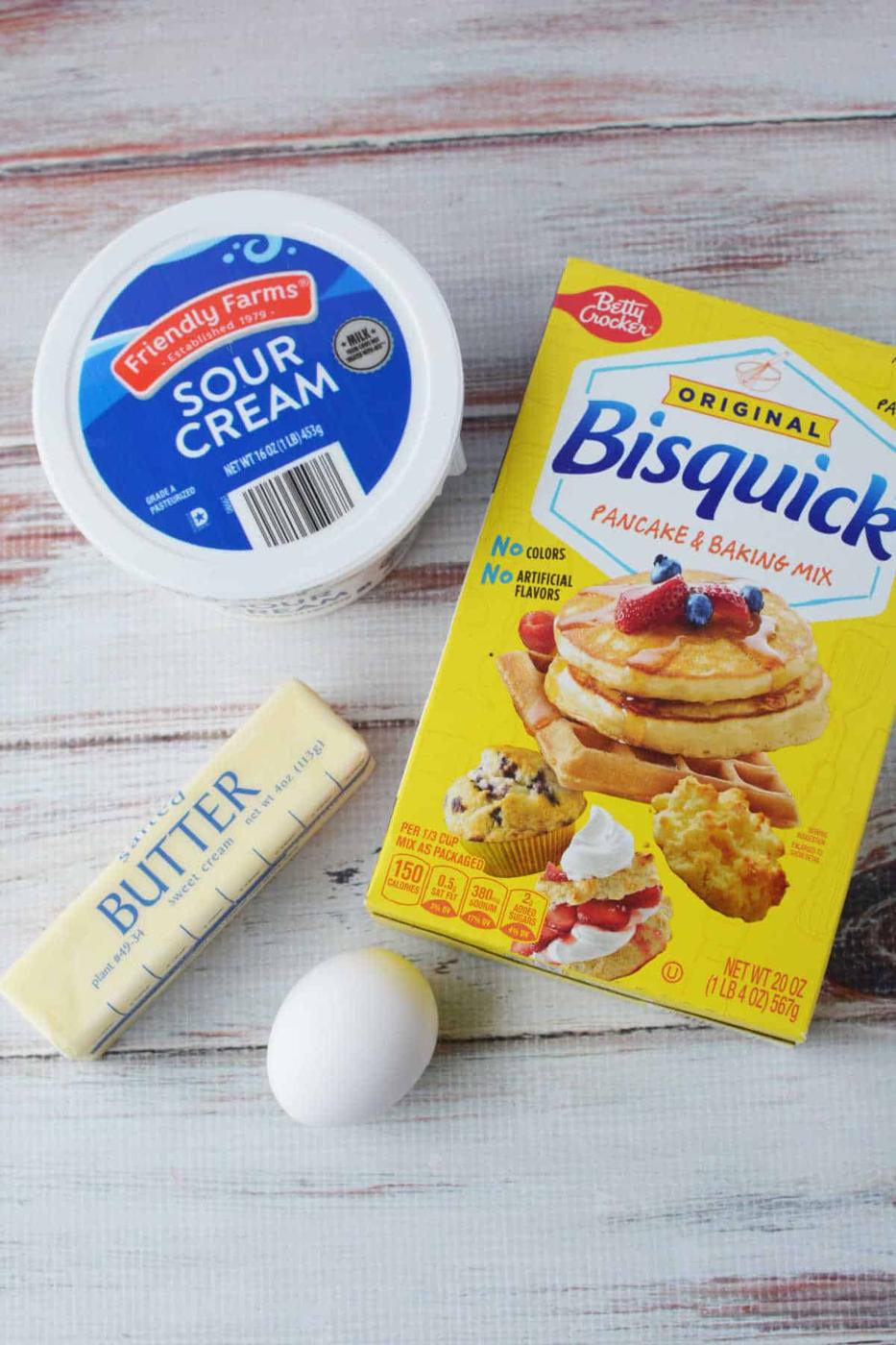 Ingredients for delicious Sour Cream Biscuits: a box of Bisquick, tub of sour cream, stick of butter, and an egg on a wooden surface.