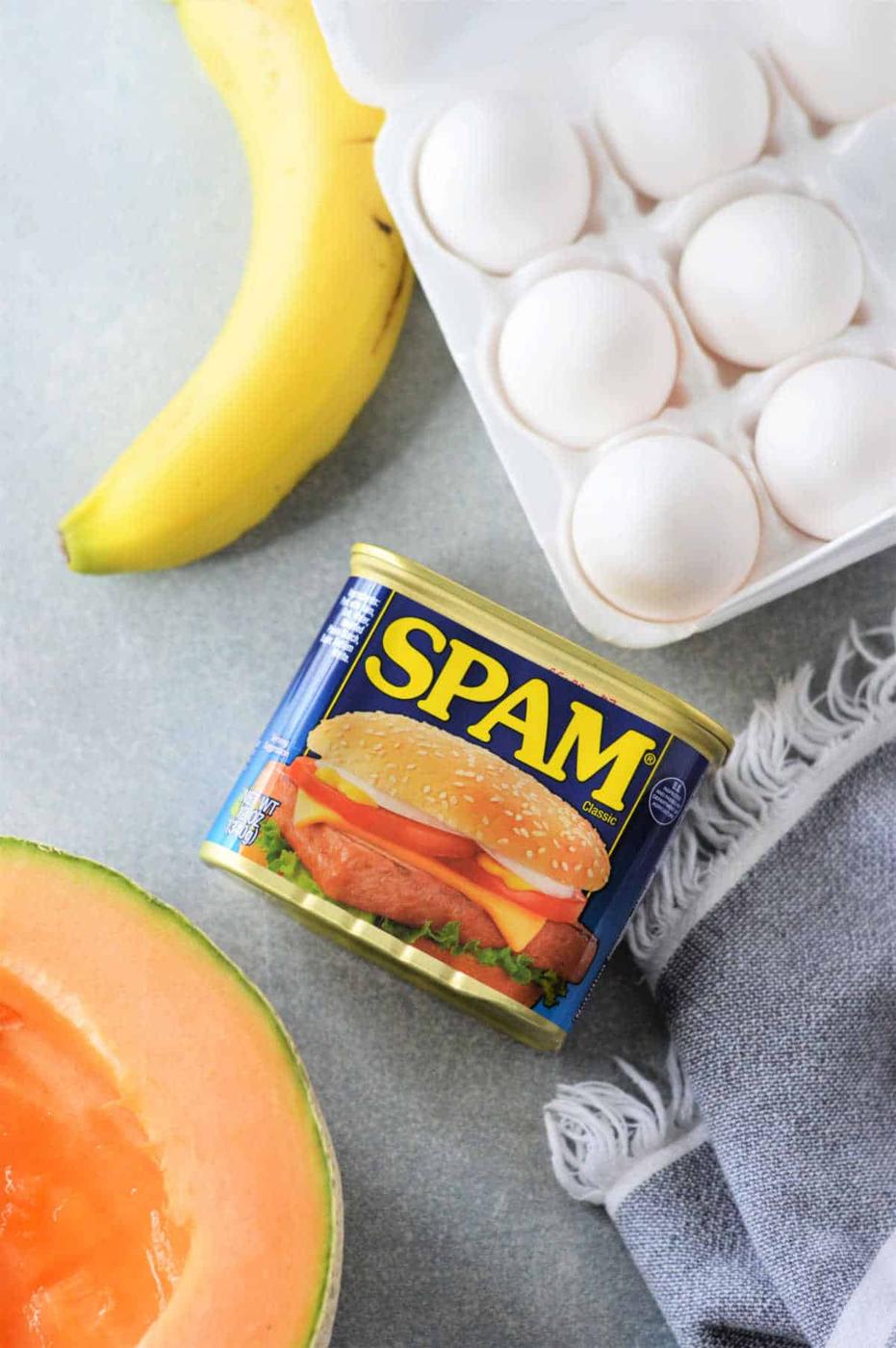 A can of Spam, a carton of eggs ready for a delicious Spam and Eggs recipe, a ripe banana, half a cantaloupe, and a gray cloth rest on the countertop.