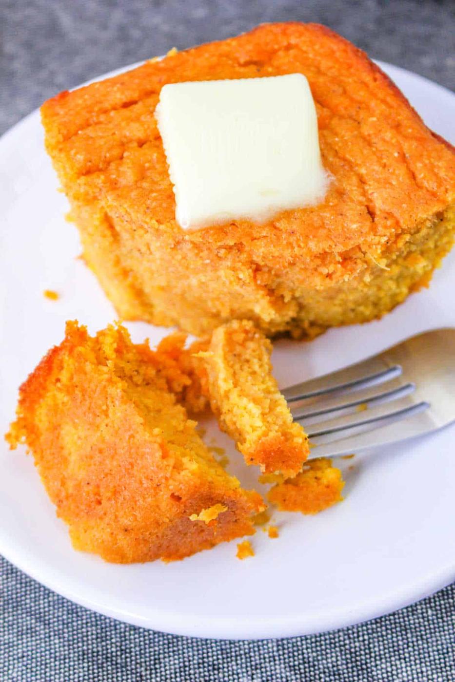 A slice of delicious spoonbread recipe, sits on a white plate with a pat of butter melting on top and a fork beside it.
