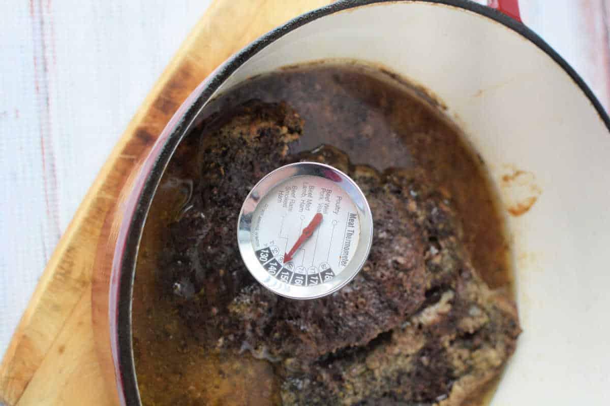A succulent Stove Top Pot Roast sits in a pot on a wooden surface, with a thermometer revealing its perfect temperature of 150 degrees Fahrenheit.