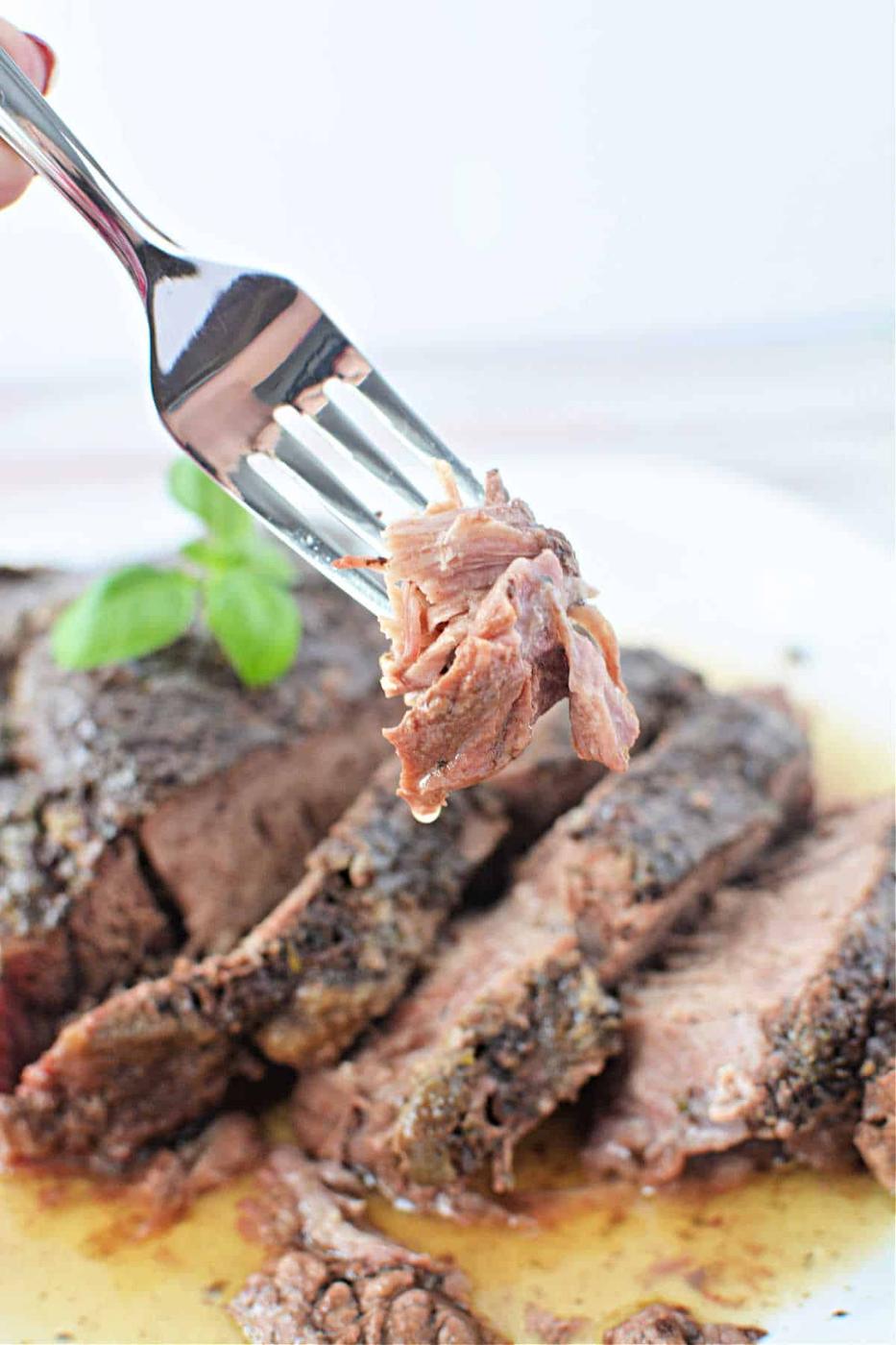 A fork holds a piece of sliced, cooked Stove Top Pot Roast above a plate of beef, garnished with a basil leaf.