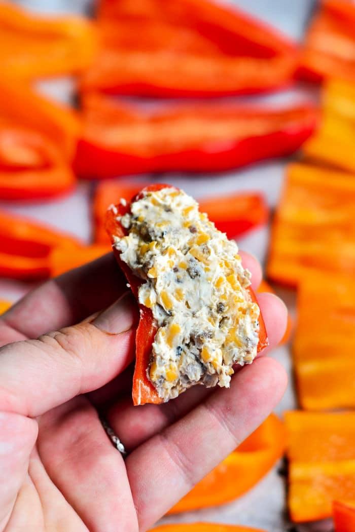 A hand holds a stuffed mini sweet pepper filled with a blend of cream cheese and herbs. Slices of red and orange peppers are blurred in the background, adding a colorful touch.