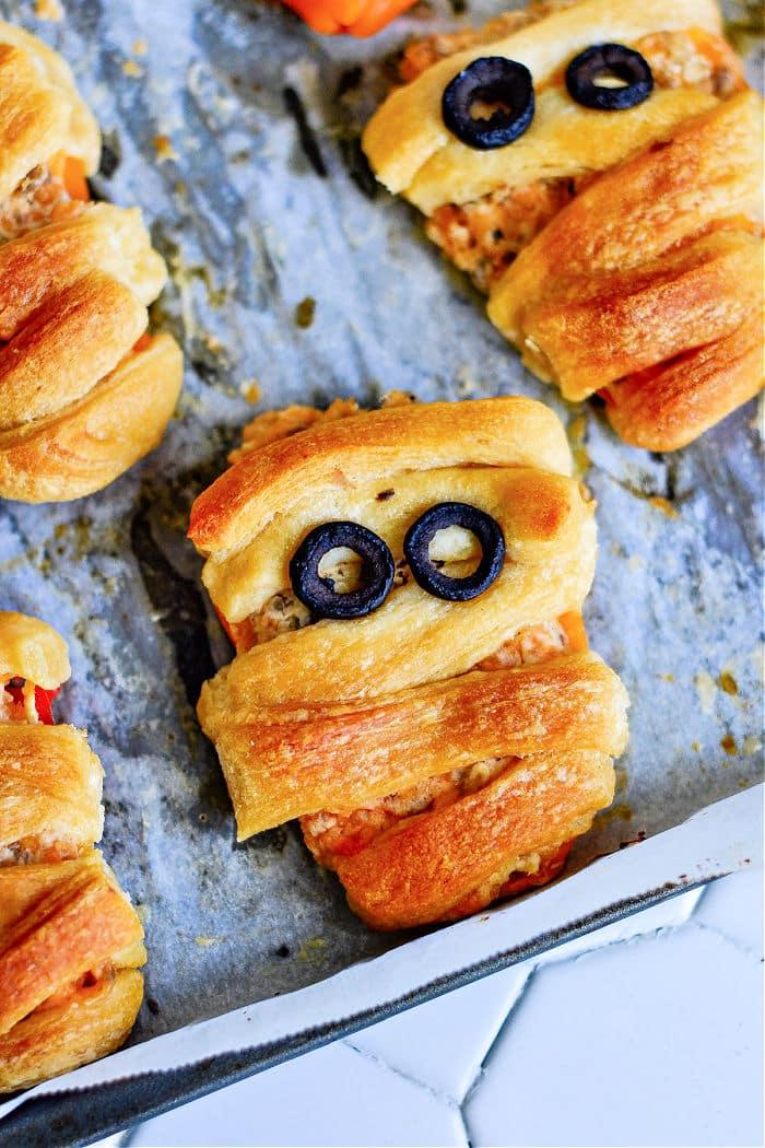 Freshly baked crescent roll mummies and stuffed mini sweet peppers rest on a parchment-lined baking sheet, the mummies shaped with olive slice eyes and wrapped dough for that classic bandage look.