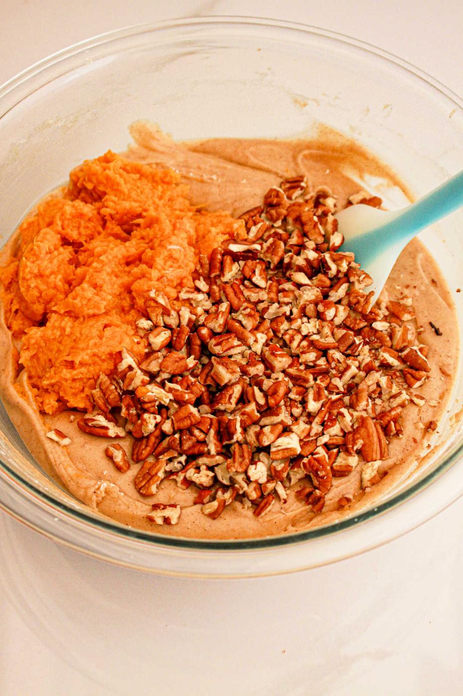 A glass bowl contains a mixture of ingredients being combined: creamy batter, orange sweet potato puree, and chopped pecans. A blue spatula is resting in the bowl, ready for mixing what will become delicious Sweet Potato Bread with Cake Mix.