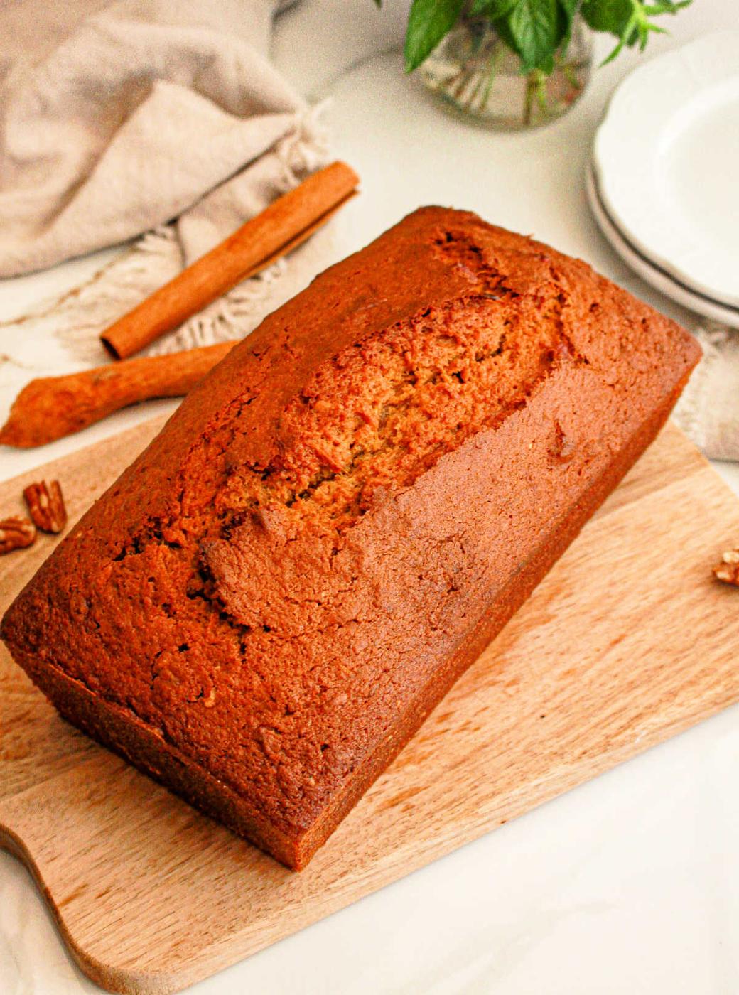 A loaf of freshly baked sweet potato bread with cake mix sits on a wooden cutting board. A cinnamon stick and some whole pecans are visible in the background, alongside a folded beige cloth and a stack of white plates.