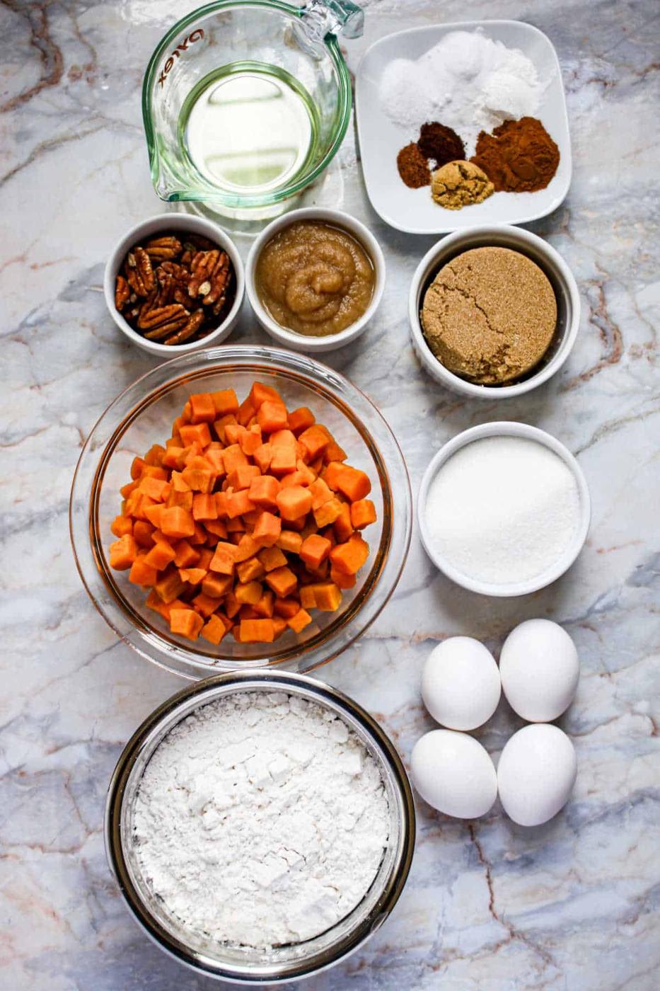 Baking ingredients on a marble surface for Sweet Potato Cake: eggs, flour, sugar, sweet potatoes, pecans, oil, applesauce, and spices.