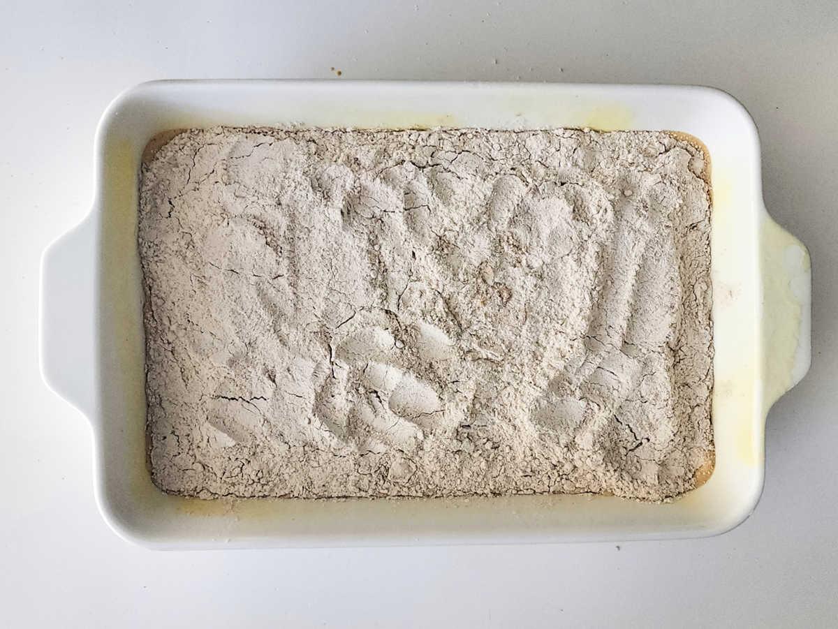 A white baking dish with a layer of flour and handprints visible on the surface, ready to become the base for a delicious Sweet Potato Dump Cake.