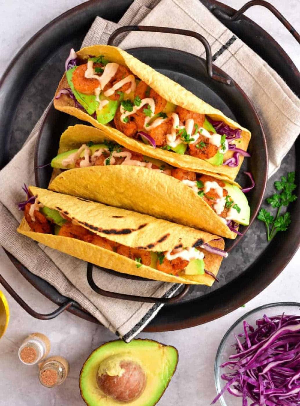 Three tacos with chicken, avocado, cabbage, and sauce sit invitingly on a plate. Beside them is a fresh avocado half and vibrant purple cabbage. These delicious tacos feature perfectly crispy taco shells made in an air fryer for the ultimate crunch experience.