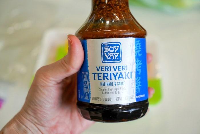 A person holds a bottle of Soy Vay Veri Veri Teriyaki marinade and sauce, perfect for Traeger smoked chicken thighs. The label is blue with white text, and the bottle cap is visible at the top. The background appears to be out of focus.