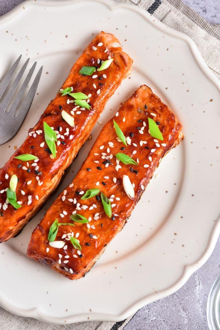 Teriyaki salmon air fried to perfection, sprinkled with sesame seeds and green onions, elegantly presented on a white plate alongside a fork.
