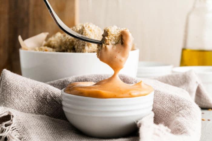 A fork dips breaded tofu nuggets into a creamy sauce in a small white bowl on a cloth, with a bowl of food in the background.