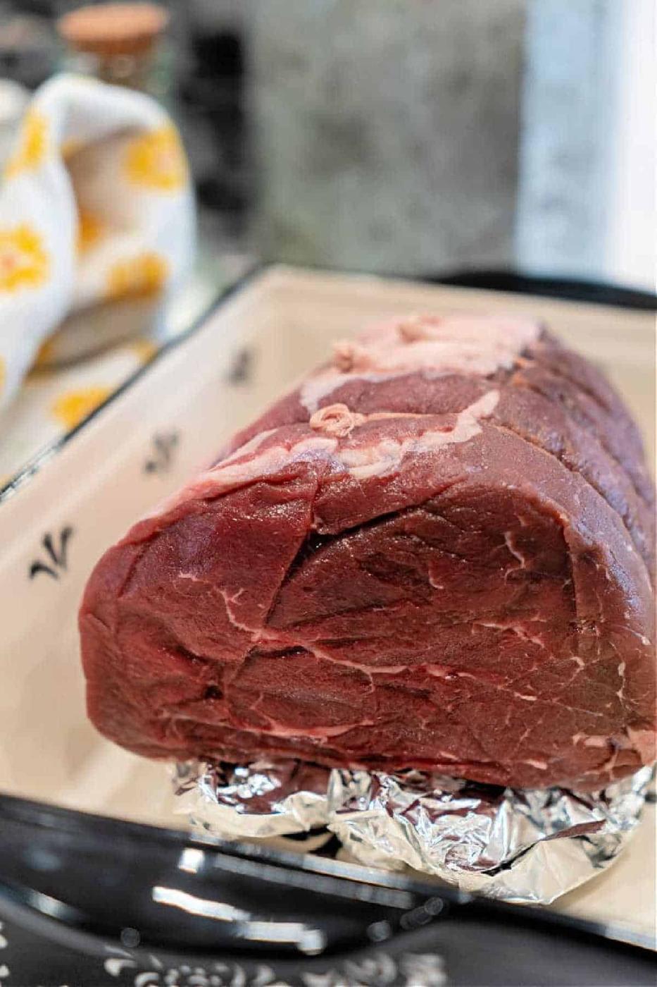 A raw top round roast sits on foil in a ceramic baking dish, ready to be cooked—perfect for your next Top Round Roast Recipe.