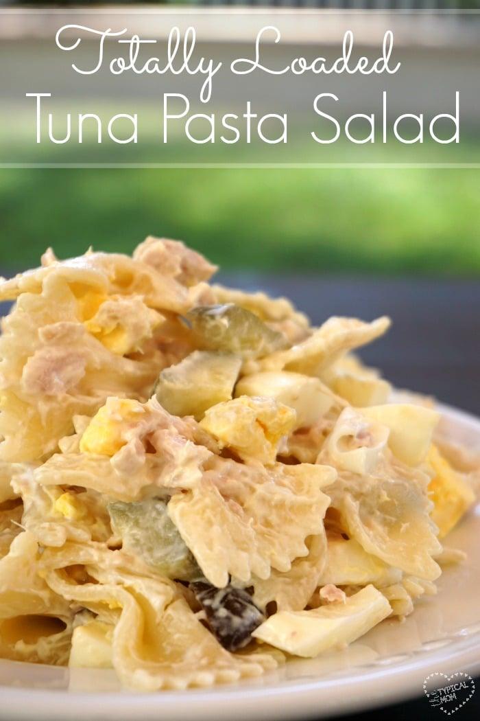 Close-up of a serving of tuna pasta salad with bow-tie pasta, chunks of tuna, diced pickles, and pieces of boiled egg. The blurred background highlights text at the top reading Totally Loaded Tuna Macaroni Salad Recipe.