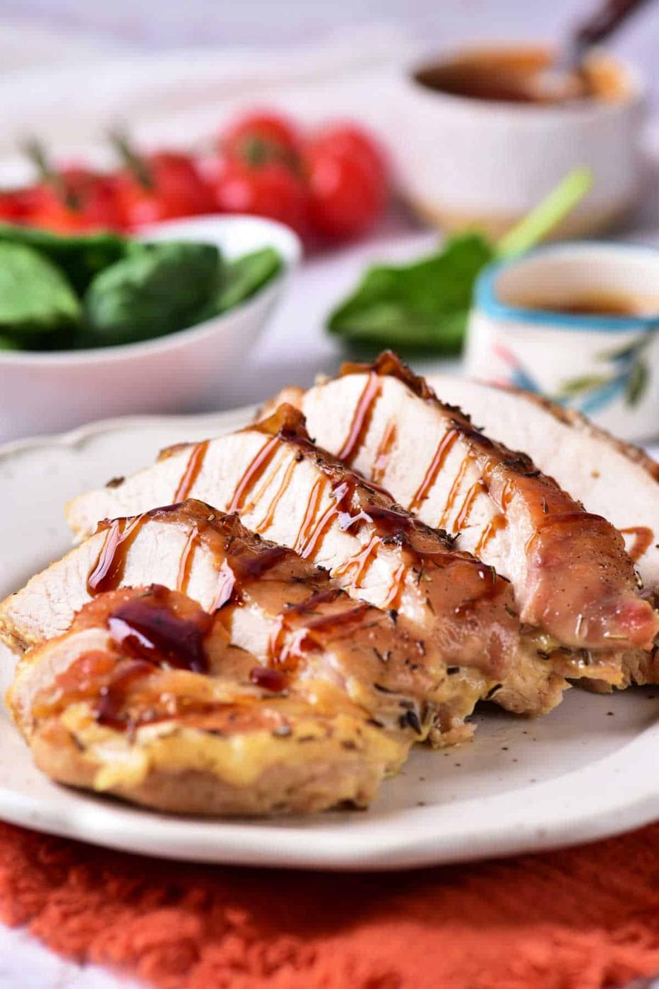 A white plate with slices of cooked, seasoned turkey breast drizzled with sauce. The plate rests on an orange cloth. In the background, there is a bowl of spinach leaves, a bunch of tomatoes, and a cup of sauce.