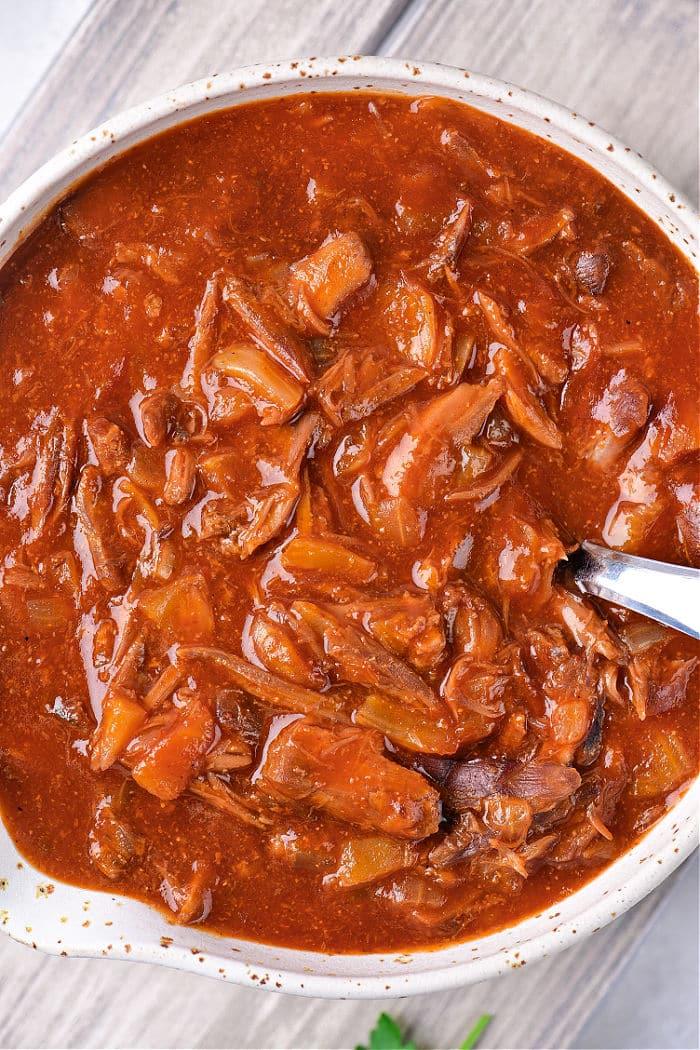 A bowl of rich gravy with turkey neck gravy, featuring tender shredded meat and vegetables, served with a silver spoon.