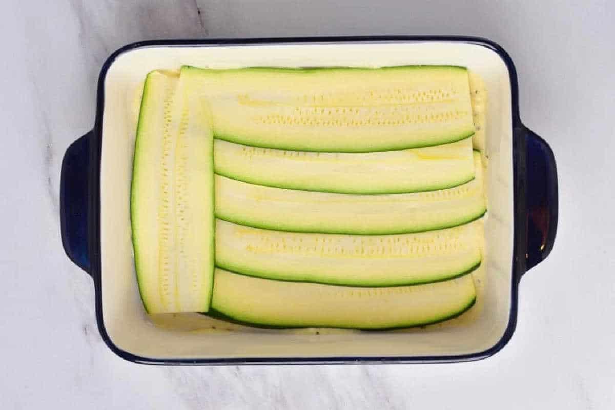 Rectangular dish with thin, raw zucchini slices neatly layered inside, ready to become a delicious vegetable lasagna with white sauce.
