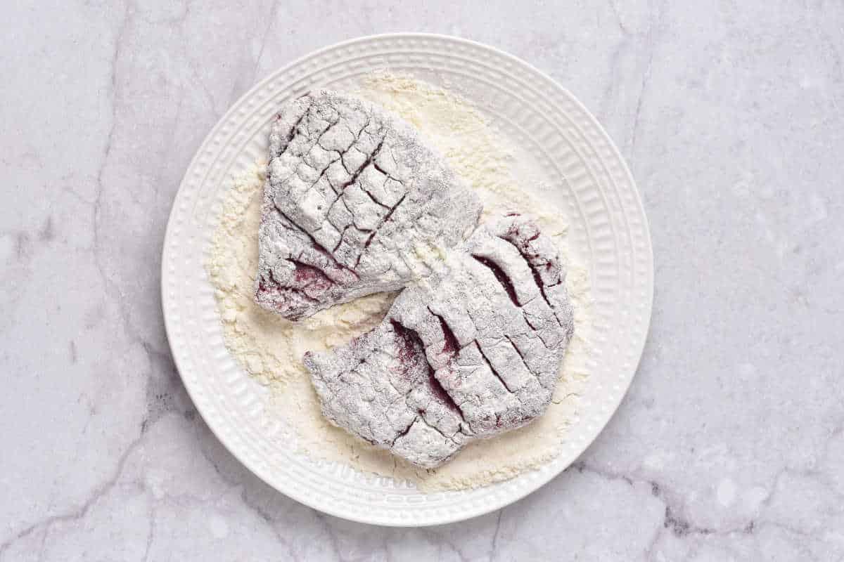 Two raw, flour-dusted deer steak rest on a white plate, placed on a marble surface
