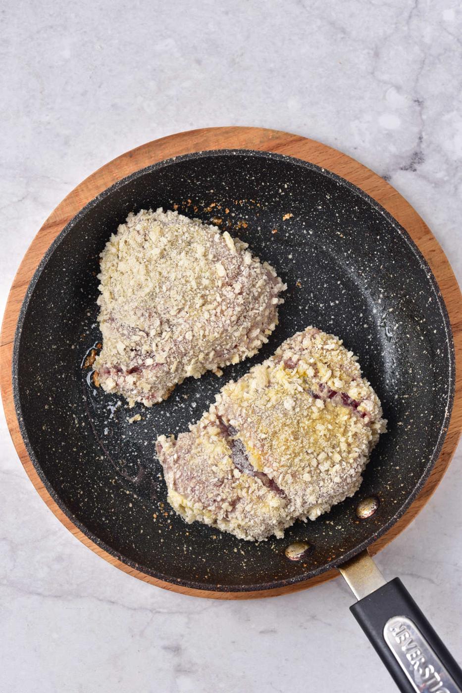 Two breaded venison steaks are sizzling in a non-stick frying pan on a marble surface in process for crafting a perfect venison cube steak recipe.