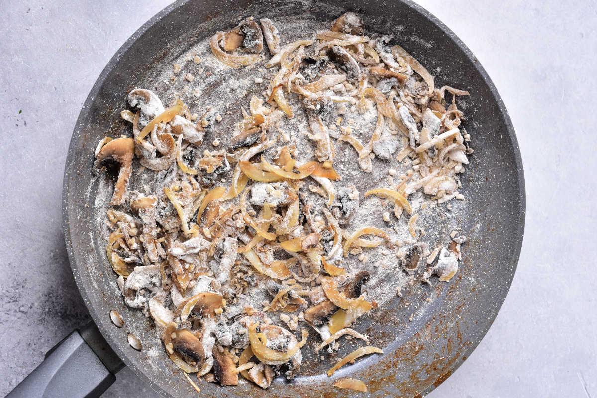 Sliced mushrooms and onions cooking with flour in a large skillet—an essential step in preparing hearty Venison Stroganoff—on a light countertop.